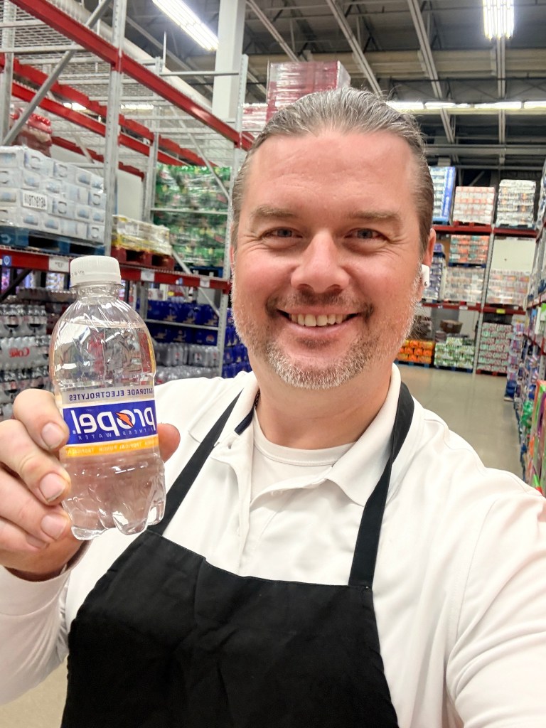 A smiling man in an apron holds a bottle of Propel water in a retail store aisle, surrounded by shelves stocked with drinks.