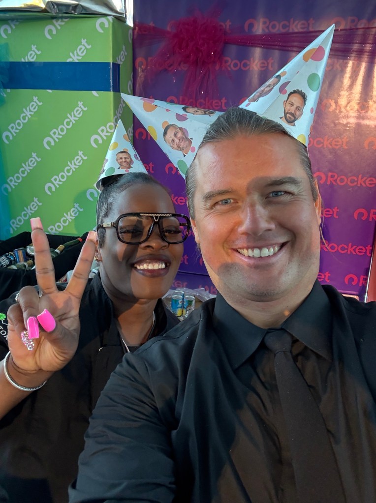 Two people wearing festive party hats pose for a selfie, one making a peace sign. They are in front of a colorful backdrop with wrapped gifts.