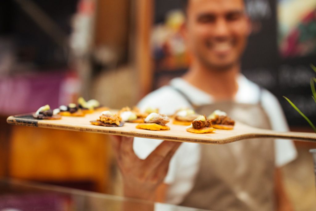 A smiling person holding a wooden tray with a variety of small gourmet appetizers.